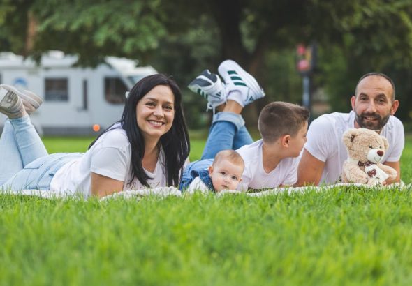 Happy family lying on grass with RV in the background, enjoying a sunny outdoor picnic.